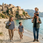 Family of four walking along a sandy beach with a vendor cart of fruit beside a signpost and a village on the rocky coast in the background
