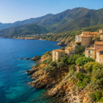 Coastal village with stone houses perched on a rocky cliff overlooking clear blue sea, with green vegetation and distant mountains in the background.
