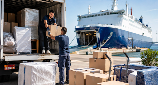 Two workers unload a cardboard box from a truck at a port with a large blue-and-white ferry in the background.